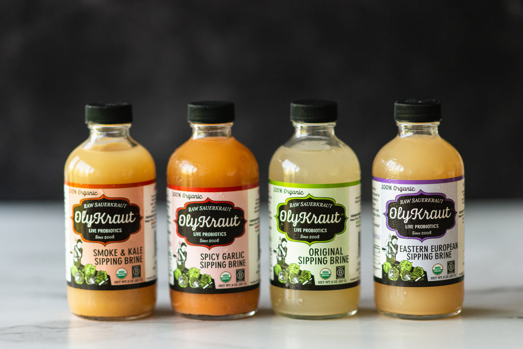 A lineup of four 8 ounce bottles of Sipping Brine from OlyKraut - Smoke & Kale, Spicy Garlic, Original, and Eastern European. 